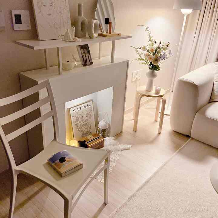 暖かいベージュと白の色調のリビング、暖炉コンソールと白いチェア、花瓶が置かれたスツールが調和する居心地の良い空間