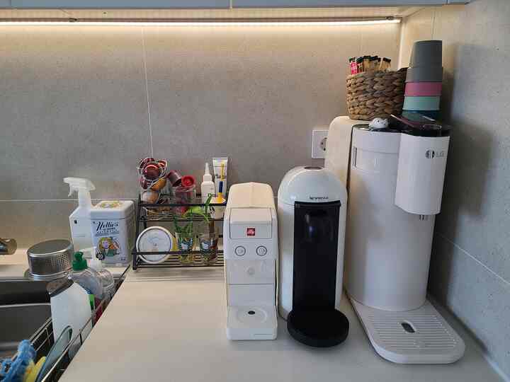 Beige-toned kitchen countertop featuring three coffee machines, cleaning supplies, and a clock in a tidy home cafe setting