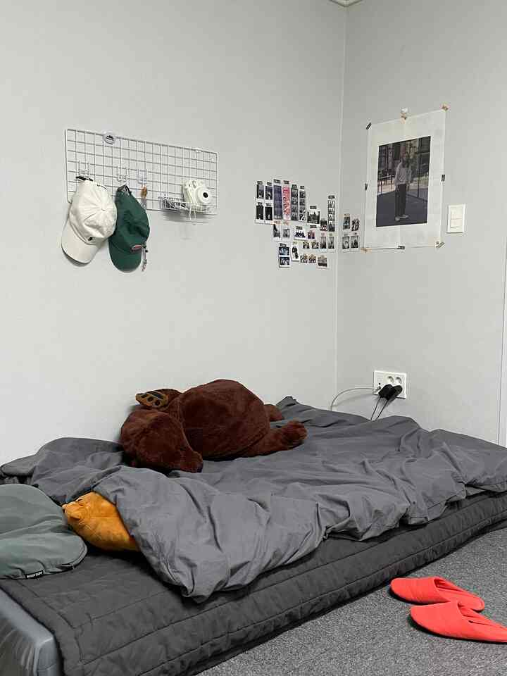 グレーのトーンのワンルーム寝室、床に置かれたマットレスと掛け布団、そしてブラウンのクマのぬいぐるみが配置された居心地の良い空間