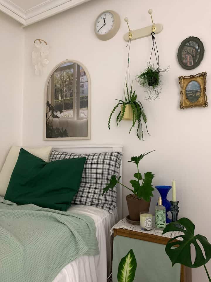 White and green toned bedroom featuring a bed with green pillows, nightstand, hanging plants, and vintage picture frames creating a cozy atmosphere