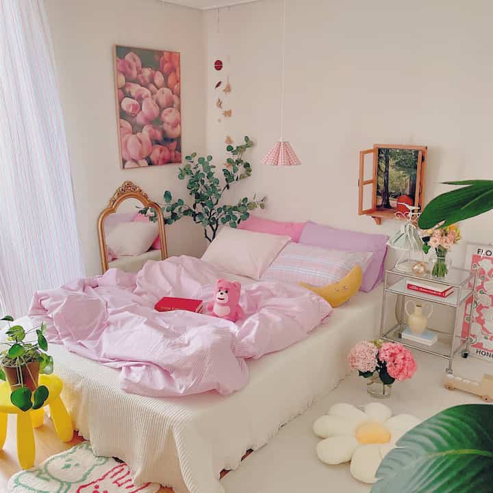 Pink and white toned bedroom featuring a large central bed with plush cushions and greenery, creating a cozy atmosphere
