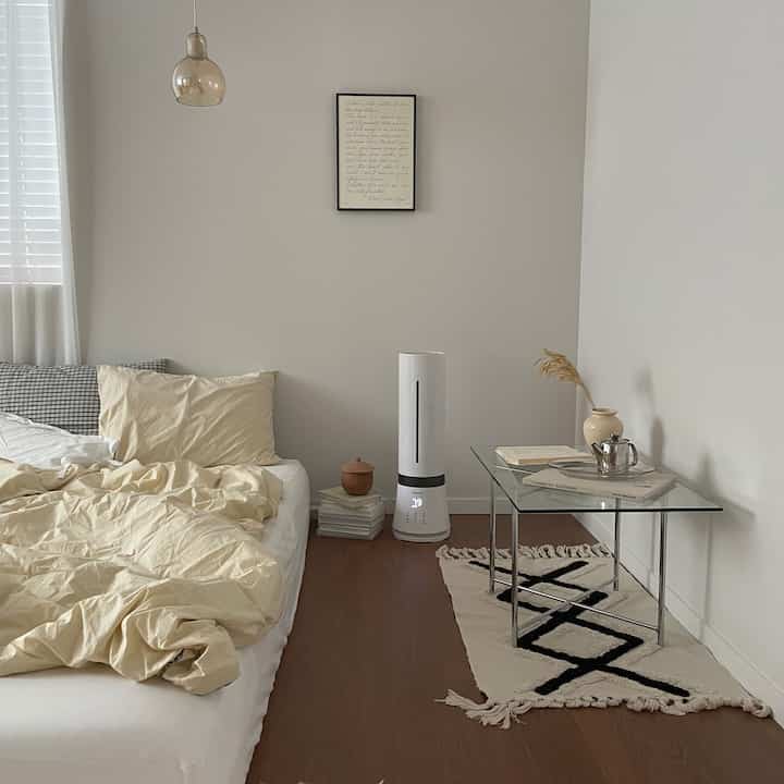 Bright white-walled bedroom with natural wood-tone flooring, cream bedding, and a transparent glass table in a simple setting