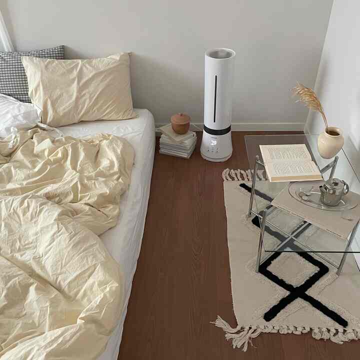Natural beige-toned bedroom featuring cream bedding, wooden floor, glass table, and patterned rug in a minimal setting