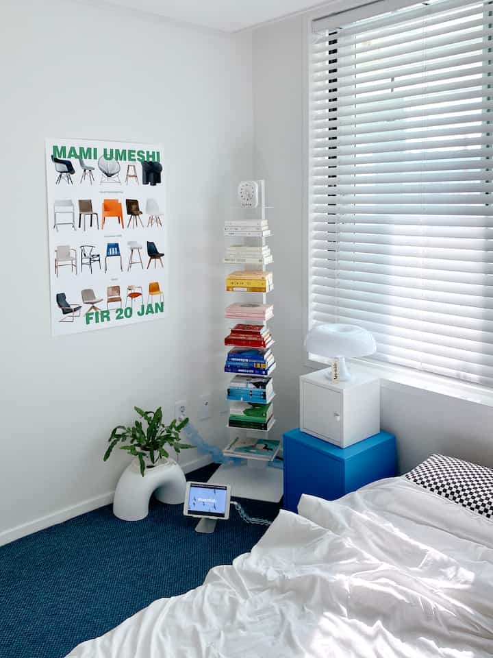 A modern bedroom in white and blue tones featuring a poster on the wall, color-organized vertical bookshelf, and horizontal blinds on the window, creating a clean vibe