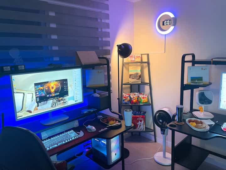 Compact game room with blue LED lighting and black furniture, featuring two desks and various accessories