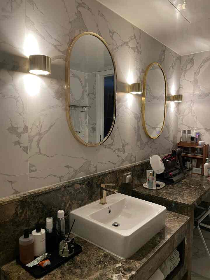 Hotel-like bathroom with gold-framed mirrors and marble tiles, featuring a washbasin and wall sconces that create a modern ambiance