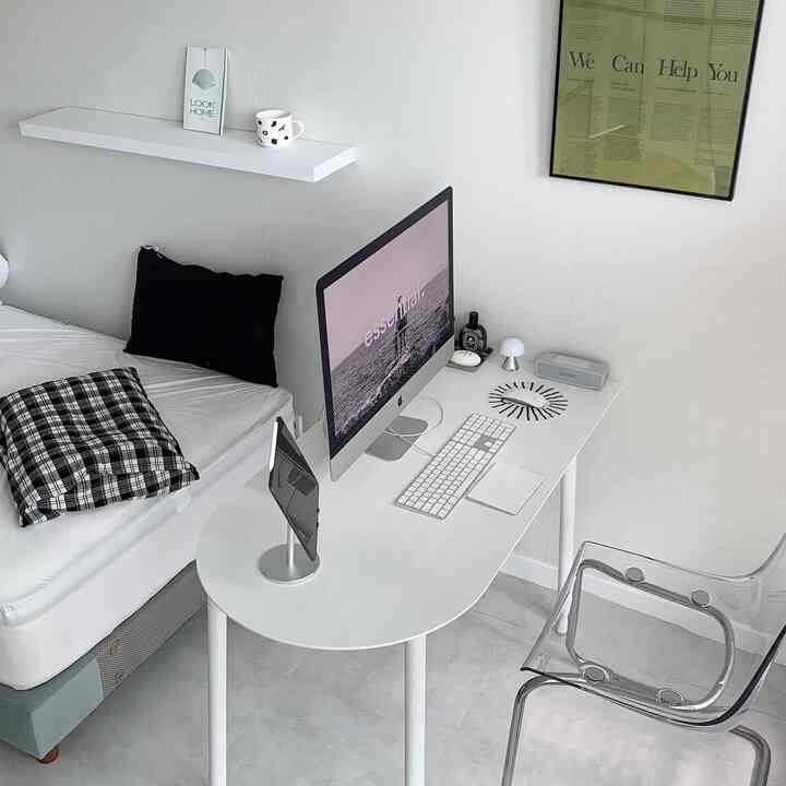 White-toned compact bedroom and home office space featuring a white desk, transparent chair, bed, and framed wall art with modern style
