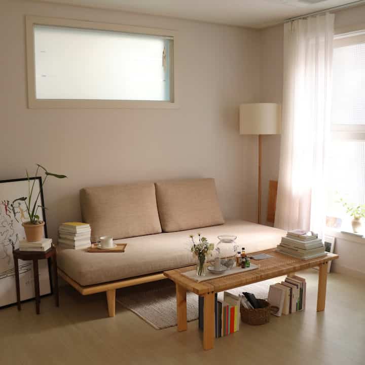 Beige and natural-toned studio living room featuring beige fabric sofa, rattan coffee table, floor lamp, and sheer curtains creating a cozy atmosphere