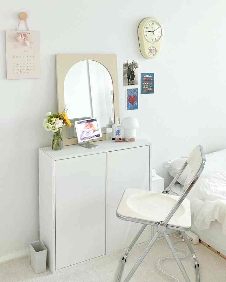 白を基調とした寝室、シンプルな収納キャビネットとアーチ型の鏡、レトロな時計、折りたたみ椅子が配置された清潔感のある空間