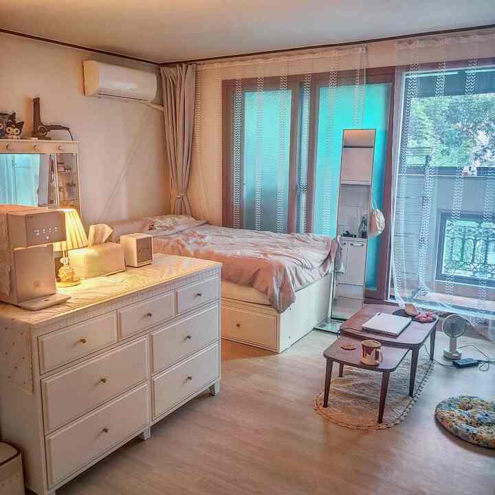 Natural-tone studio apartment bedroom featuring white bed frame, dresser, coffee table, and sheer curtains with cozy ambiance