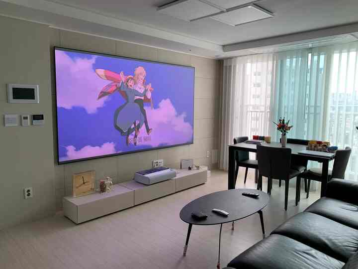 明るいホワイトトーンのリビング、大きく鮮明なプロジェクタースクリーンと黒いソファ、ダイニングテーブルが配置されたモダンな空間