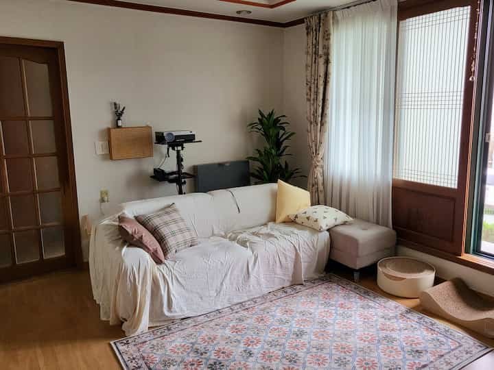 Ivory and beige toned living room featuring white sofa, patterned rug, floral curtains by the window, and cat scratching posts, creating a cozy atmosphere