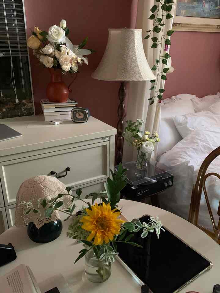 ピンクの壁にヴィンテージ調のランプとオレンジと白の花瓶が配置された寝室、丸テーブルの上にはひまわりとアイパッドが置かれた雰囲気のあるインテリア