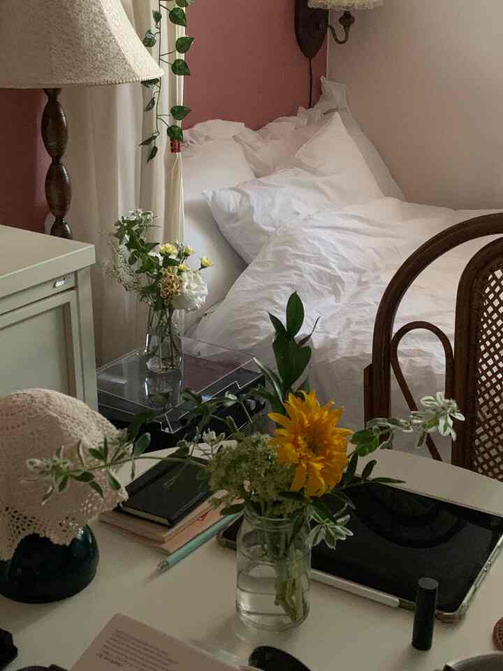 A cozy bedroom featuring a pink wall with white bedding, natural wood furniture, and plants arranged for warmth