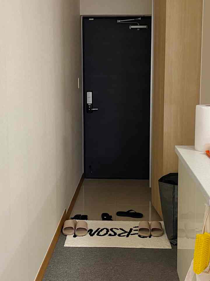 Narrow entrance space with black door, wood-toned walls, white entrance mat, and slippers arranged on the floor