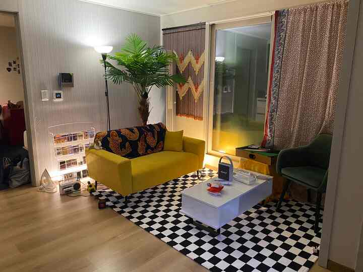 黄色のソファと大きな人工観葉植物が配置されたリビング、チェッカーボードラグと白いコーヒーテーブルがある雰囲気のあるインテリア