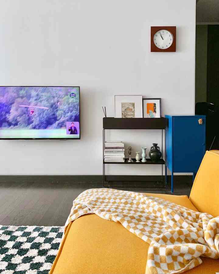 A modern living room with white walls and dark brown flooring featuring a yellow sofa with a checkered throw, black shelving, and wall-mounted TV