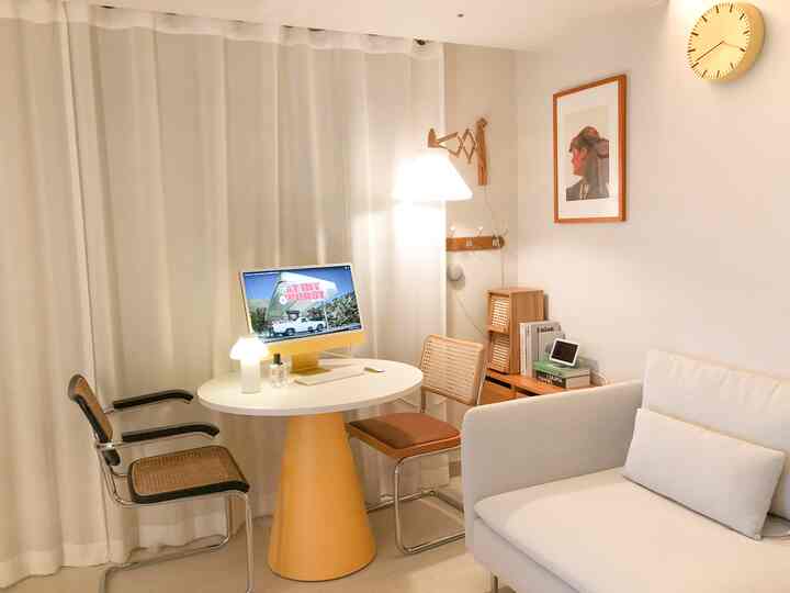 白とゴールドの色調のリビング、丸テーブルとラタンチェア、ソファ、壁時計、壁掛け照明が配置された雰囲気のあるホームオフィスインテリア