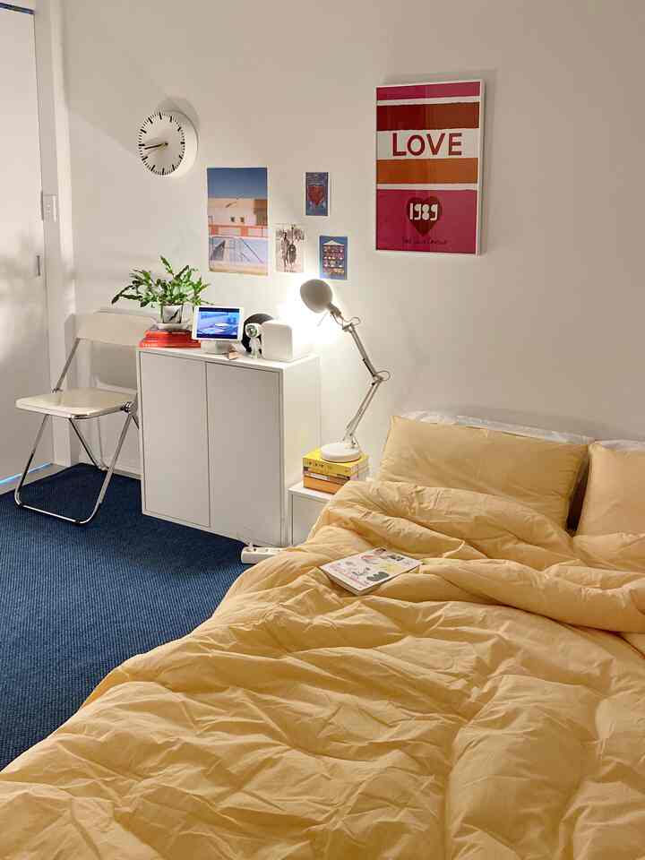 A modern bedroom with warm yellow bedding against white walls, featuring simple furniture and artistic posters creating harmony