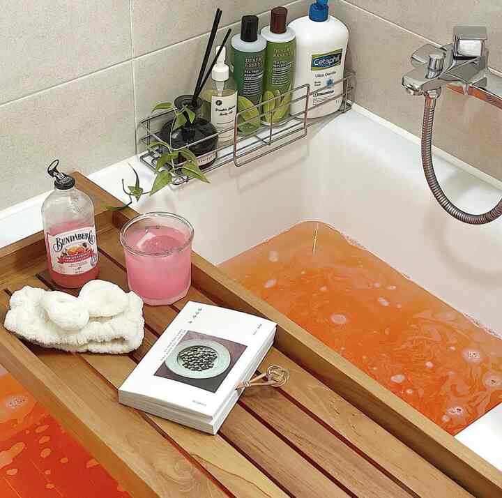 ナチュラルなベージュ色調のバスルーム、オレンジ色の水が入った浴槽と飲み物や本が置かれたバストレイのある雰囲気のあるインテリア