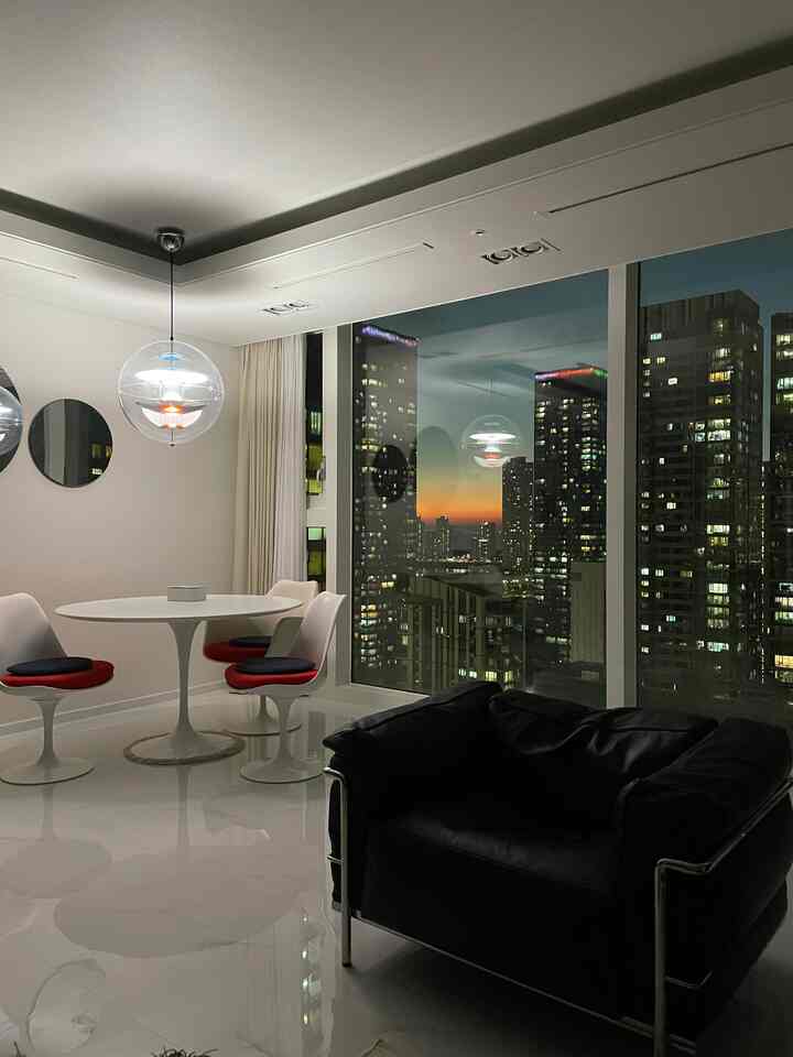 Modern dining room in white and black tones featuring a round table, chairs, pendant lights, and a large window with city nightscape and sunset view