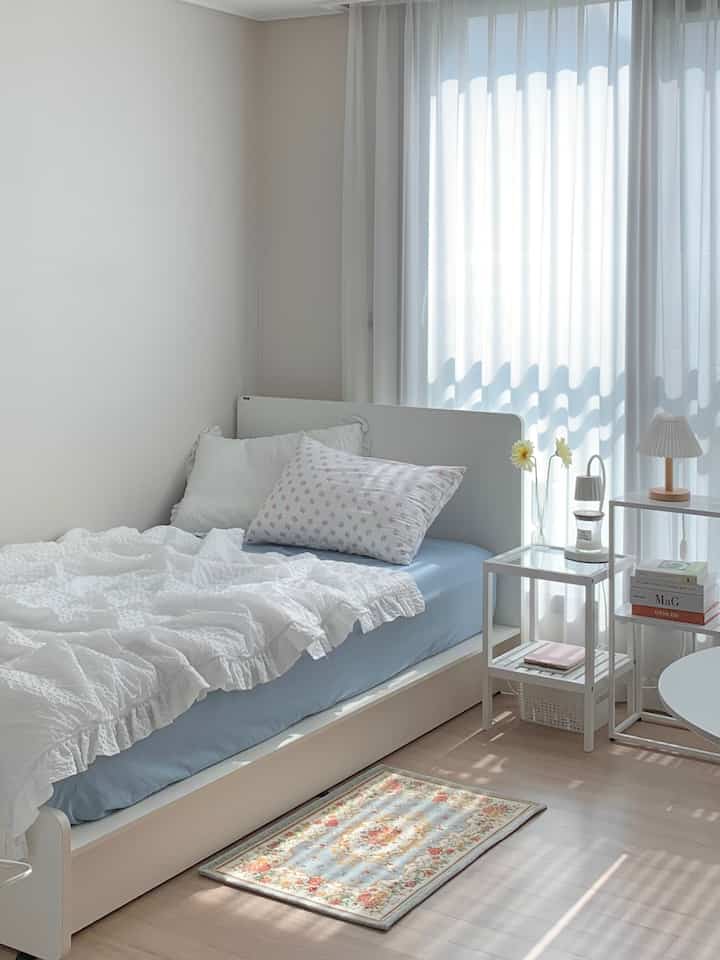 白と青のトーンでまとめられた寝室、すっきりとしたベッドや寝具、大きなシアーカーテンとナイトテーブルの照明が配置された落ち着いた雰囲気のインテリア