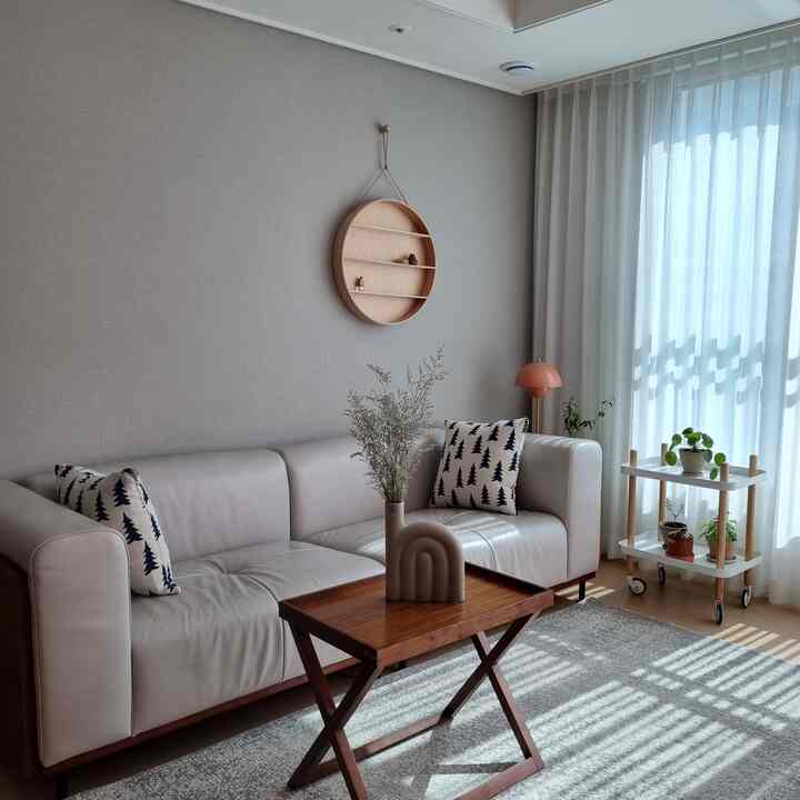 Beige and natural toned living room featuring modern sofa, wooden coffee table, large window with white curtains, and plants arranged for a cozy atmosphere