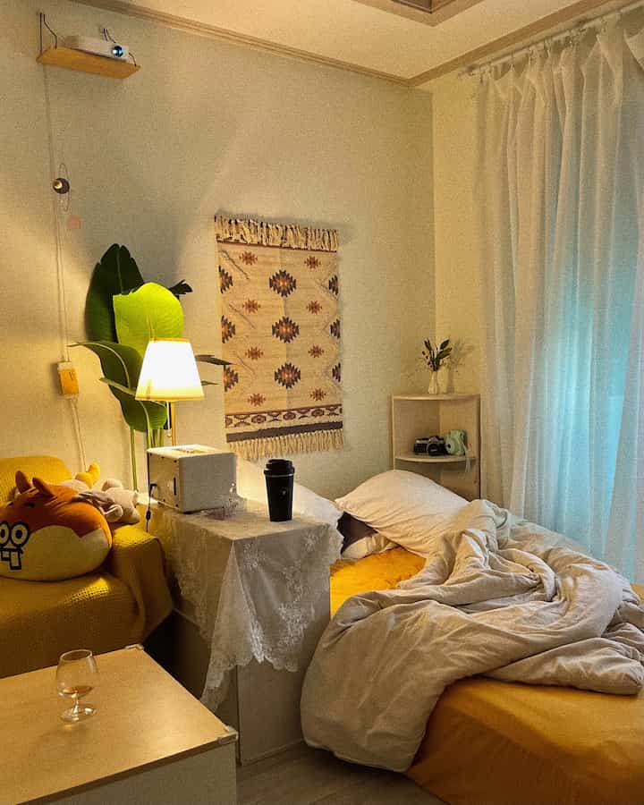 A cozy natural-toned bedroom featuring yellow bedding, white curtains, nightstand, and table lamp creating a warm atmosphere
