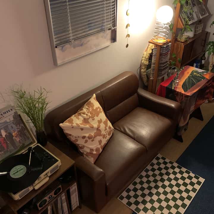 Cozy living room with wood tones on floor and brown leather sofa, blinds on window, vintage decor, and plants arranged around