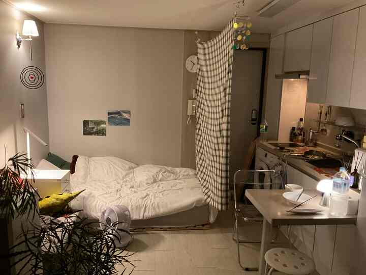 白とナチュラルトーンの5坪ワンルーム、カーテンの間仕切りで寝室とキッチンが分かれた落ち着いた一人暮らし空間