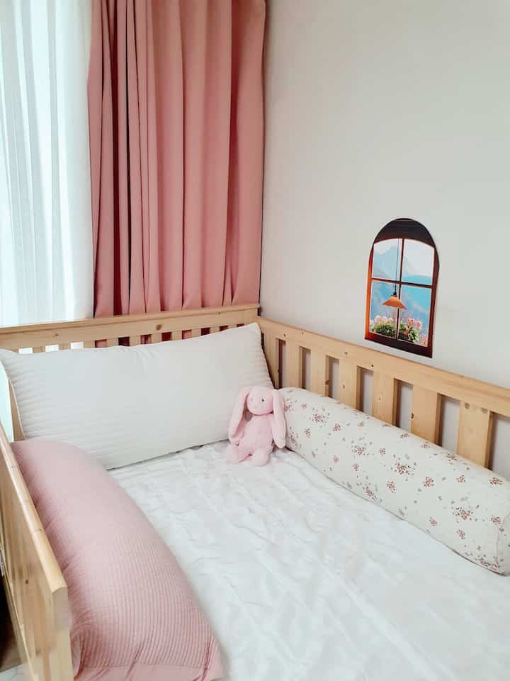 Pink-toned kids' room featuring a baby bed with cushions and an arched window poster creating a cozy atmosphere