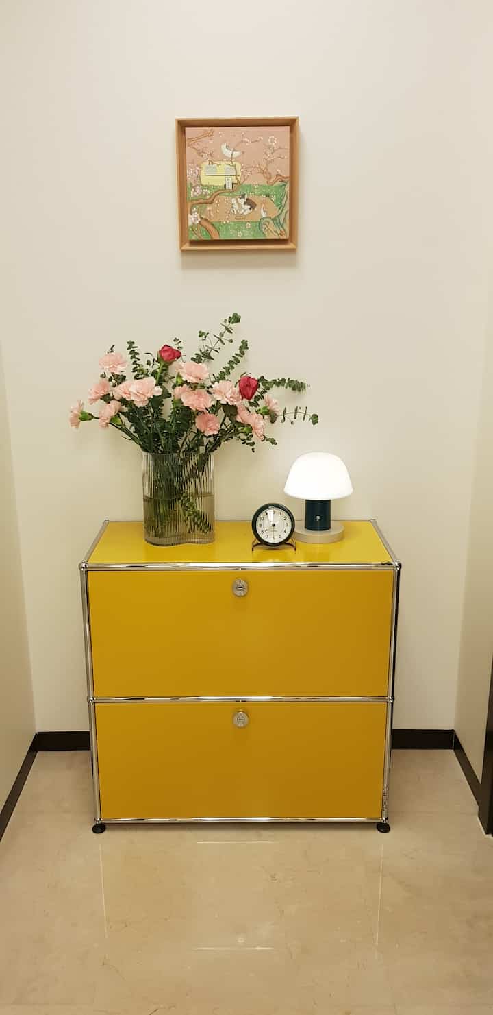 Yellow USM Haller storage topped with vase, clock, and table lamp under a wall painting in a tidy entrance space