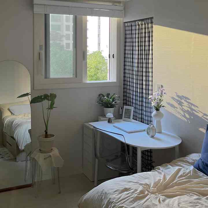 A white and natural tone studio bedroom featuring a desk, bookshelf, and a cozy, tidy home office setup
