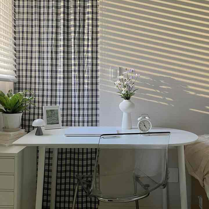White-toned home office space featuring a white desk, transparent chair, plaid curtains, and plants in a simple modern style