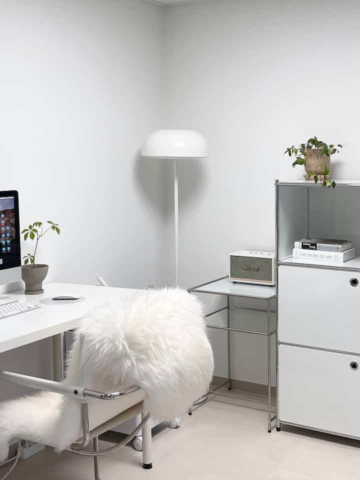 Modern study space in white tones featuring a computer desk and chair, shelving unit, floor lamp, and plants in a simple office setup