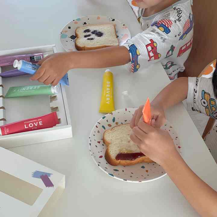White table featuring colorful plates, decorated bread, and children playing home cafe activity with assorted jam tubes