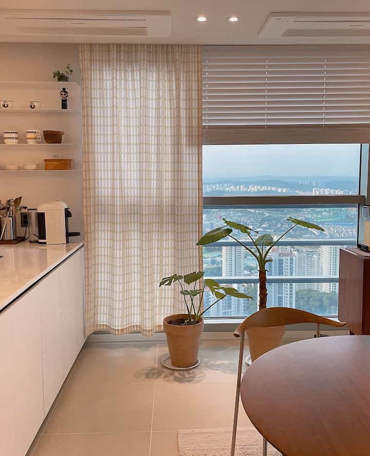 Beige and white toned kitchen space featuring a wooden dining table and chair, patterned curtain, and potted plants creating a cozy atmosphere