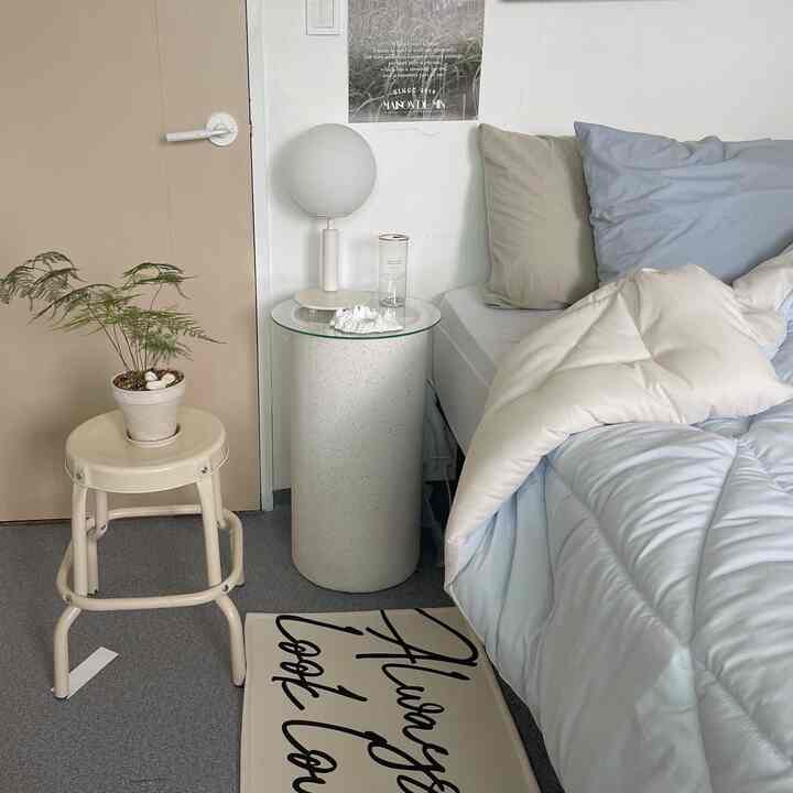 Beige and gray toned small bedroom featuring a bed with pillows and comforter, round side table with lamp, and a stool with potted plant creating a cozy atmosphere