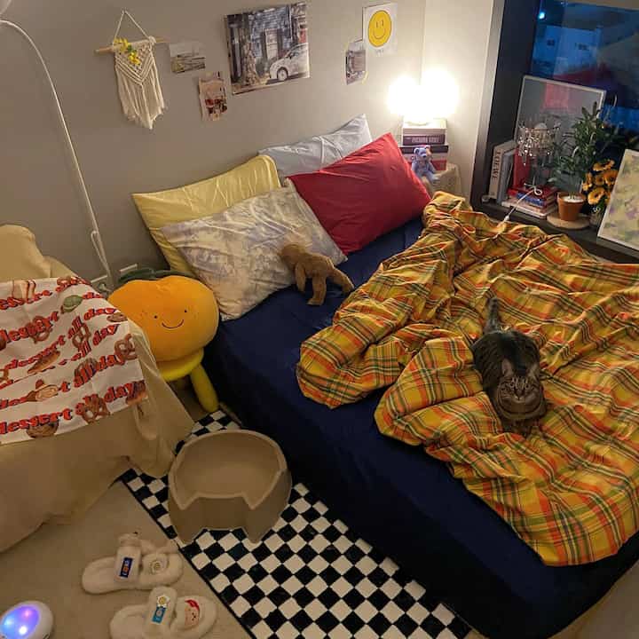 A vintage-style bedroom with yellow and red pillows and a plaid duvet, featuring a cat sitting on the bed