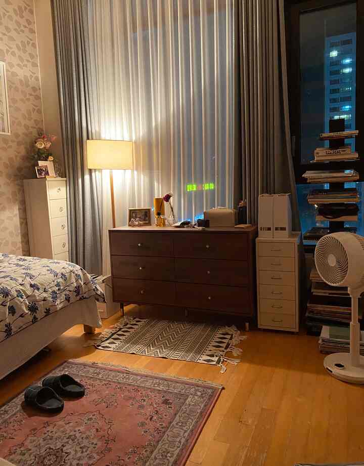 Cozy studio bedroom with warm yellow lighting, wood flooring, white curtains, featuring a bed and dresser arranged comfortably