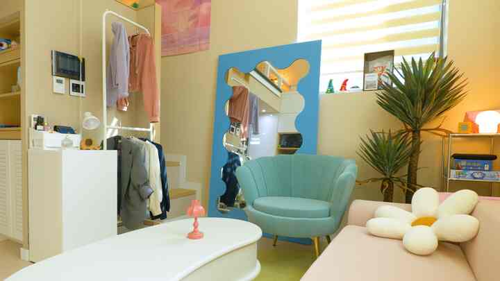 Pastel-toned cozy space featuring a pink sofa, mint green armchair, flower-shaped cushion, and a unique blue mirror in a simple living room setup