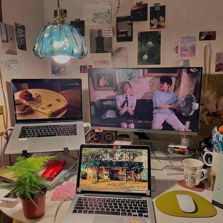 Warm beige-toned workspace featuring desk, dual laptops, large monitor, fabric wall lamp, and vintage posters on wall in home office