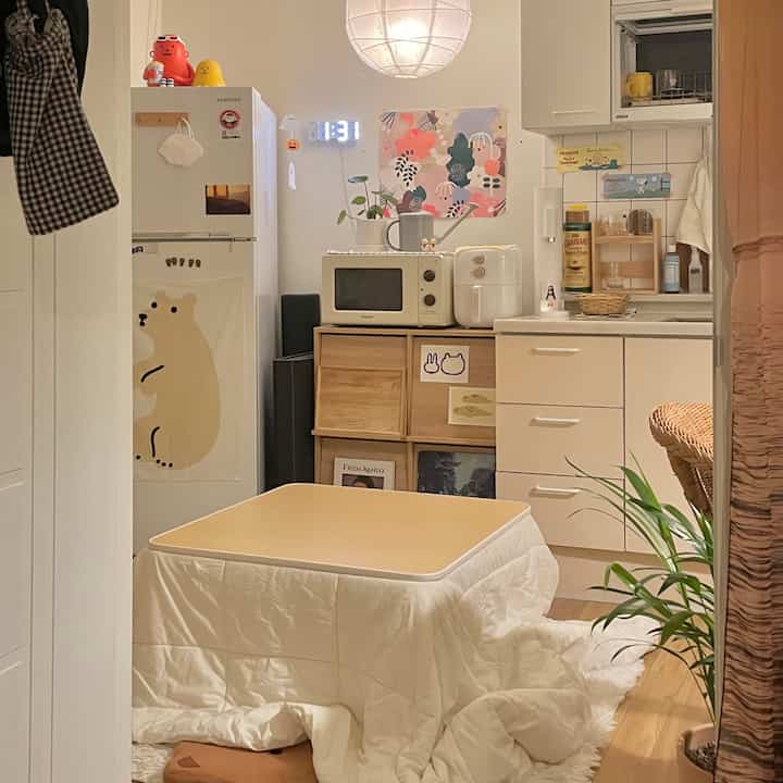 ホワイトとナチュラルブランの色調のリビング、中央にこたつテーブルと小型キッチン家電が配置された雰囲気のある空間