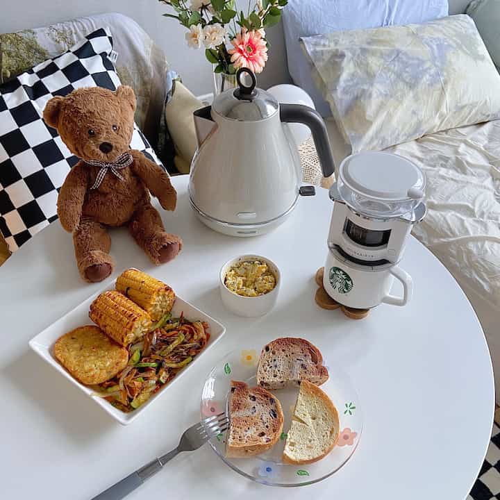 A white and beige-toned bedroom featuring a round white table with electric kettle, teddy bear, and plated breakfast creating a cozy atmosphere
