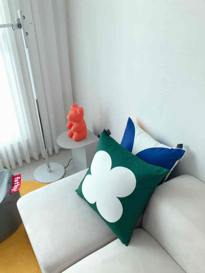 Bright white-toned living room corner featuring a white sofa with blue and green cushions, gray pouf, and small side table