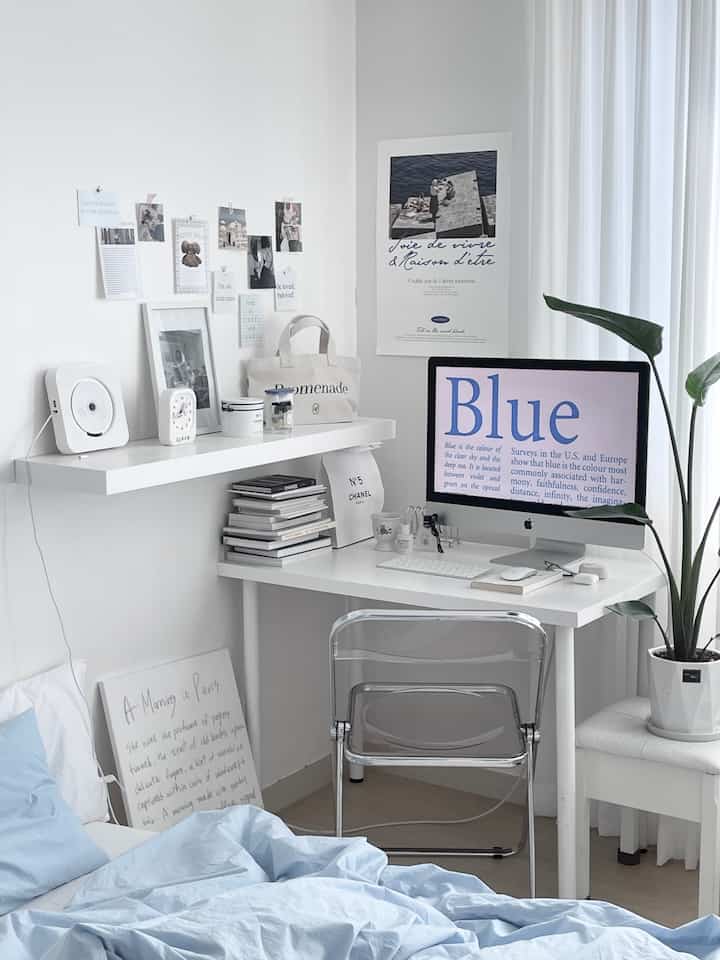 白と青のトーンが特徴の小さなホームオフィス、コンピューターデスクと透明な椅子、青い寝具が配置された落ち着いた寝室の空間