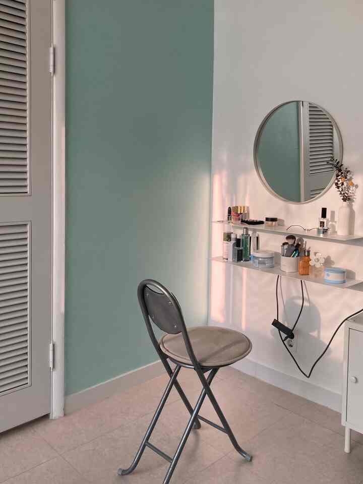 淡いミント色と白い壁のミニ化粧台空間、壁棚と丸い鏡、折りたたみ椅子が配置されたシンプルで落ち着いた雰囲気のインテリア