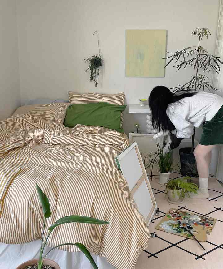 ベージュのストライプ掛け布団カバーとグリーンの枕がある寝室、複数の植物が配置されたシンプルでナチュラルな雰囲気のインテリア
