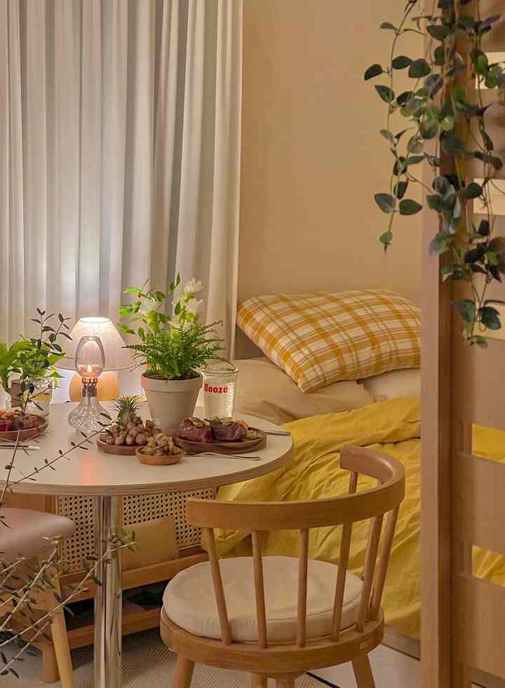 Warm beige and yellow toned studio bedroom featuring wooden dining table and chairs, curtains, and plants creating a natural atmosphere
