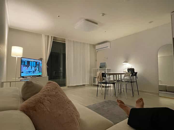 White and light brown toned living room and home office space featuring sofa, dining table, and TV in a simple modern style